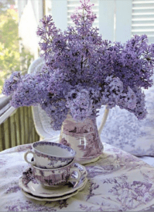 Purple lilacs in a teacup and saucer on a porch.