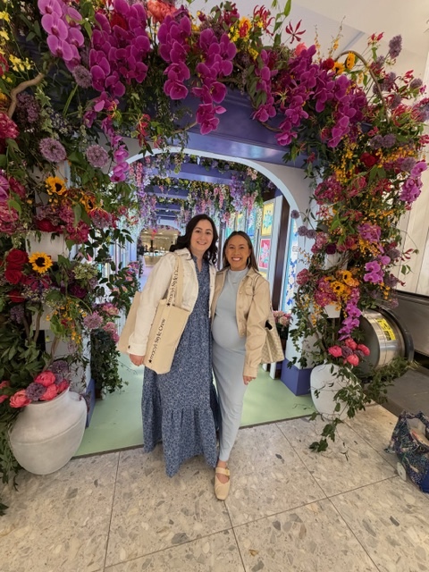 Two women pose under a vibrant indoor flower arch; one in a blue dress with a beige cardigan, the other in pale blue with a tan jacket.