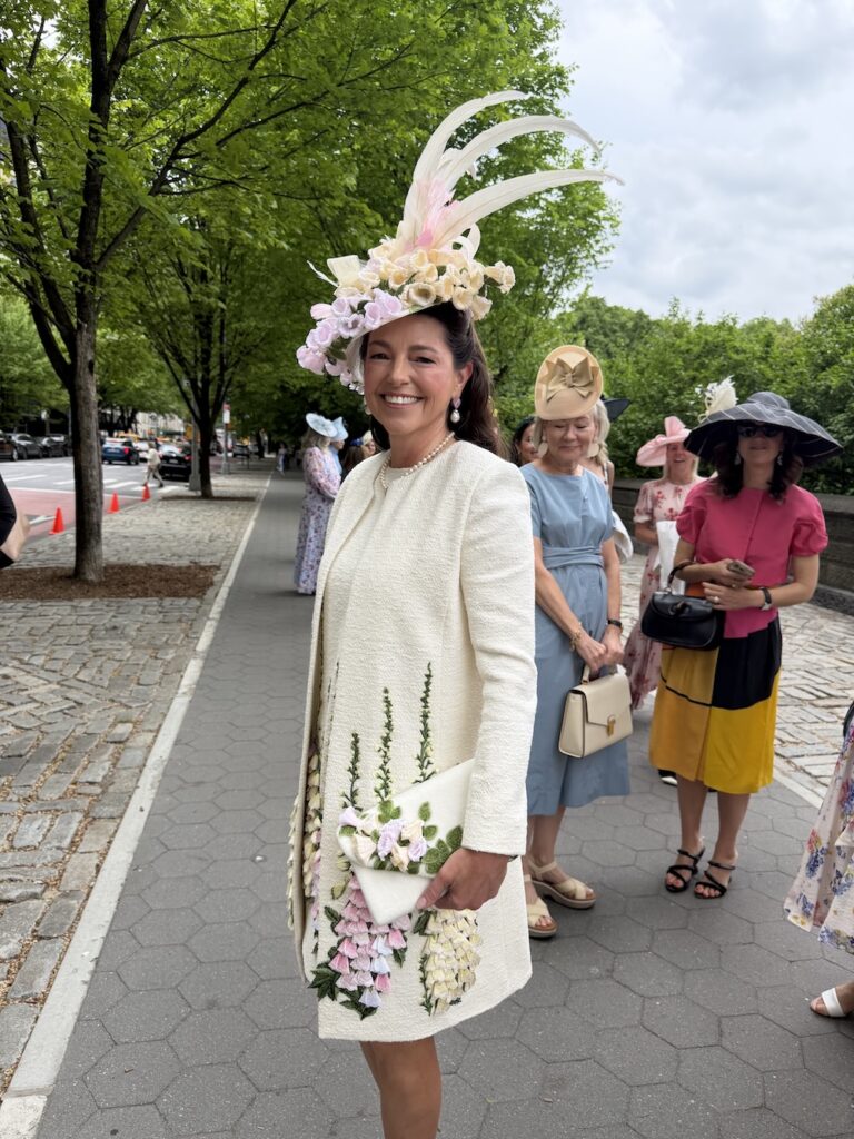 Woman wearing a cream textured coat and a large floral hat, smiling on a city sidewalk with friends in pastel outfits behind her.
