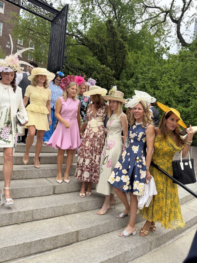 Group of elegantly dressed women wearing large hats descend stone steps at an outdoor formal event, pastel floral dresses and bright accessories visible across the line-up.