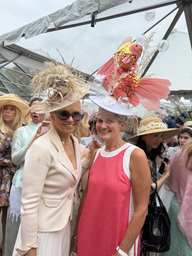 Two women at a hat parade pose together, wearing oversized decorative hats amid a crowd under a tent.