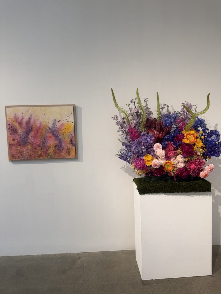 Colorful flower arrangement on a white pedestal beside a pastel abstract painting in a gallery setting.