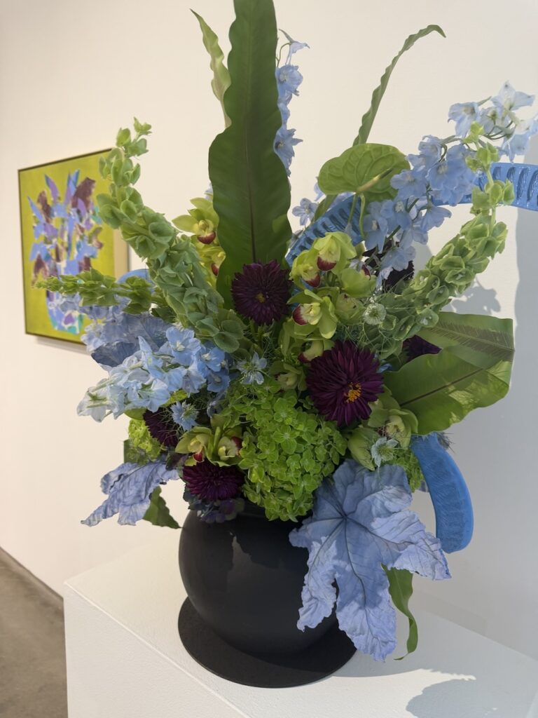 Black vase with a blue-ribbon bouquet of blue hydrangeas, delphiniums, orchids, purple dahlias, and large green leaves in an art gallery.