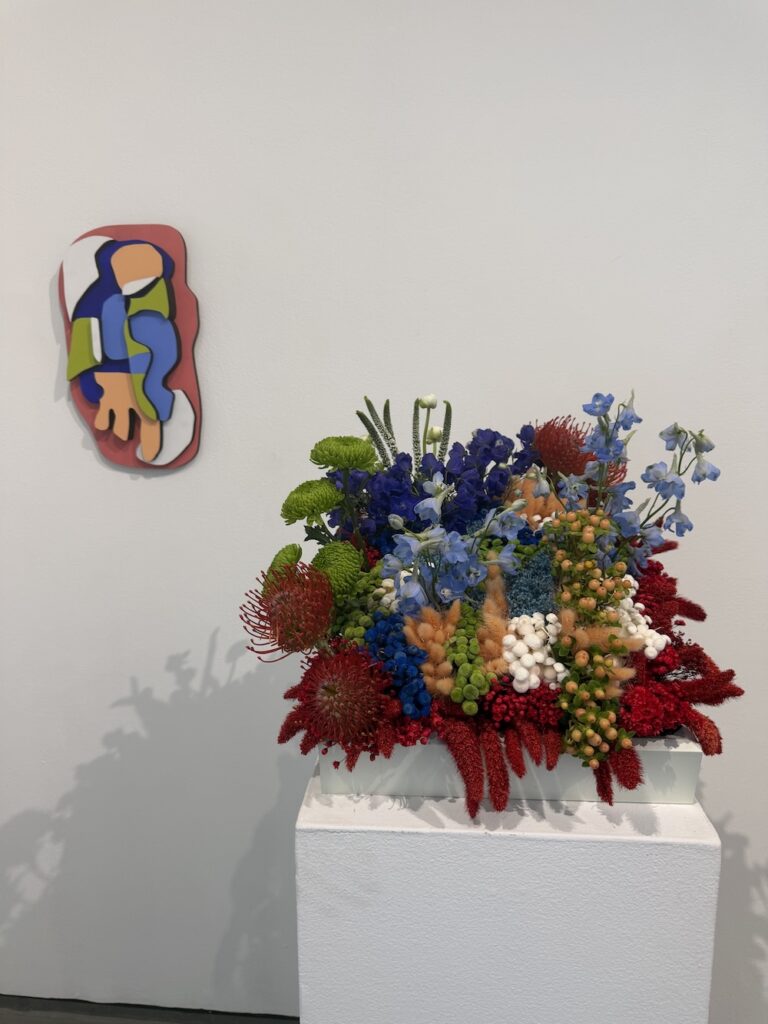 Vibrant mixed-flower arrangement in a white box on a pedestal beside a modern abstract wall sculpture in an art gallery.