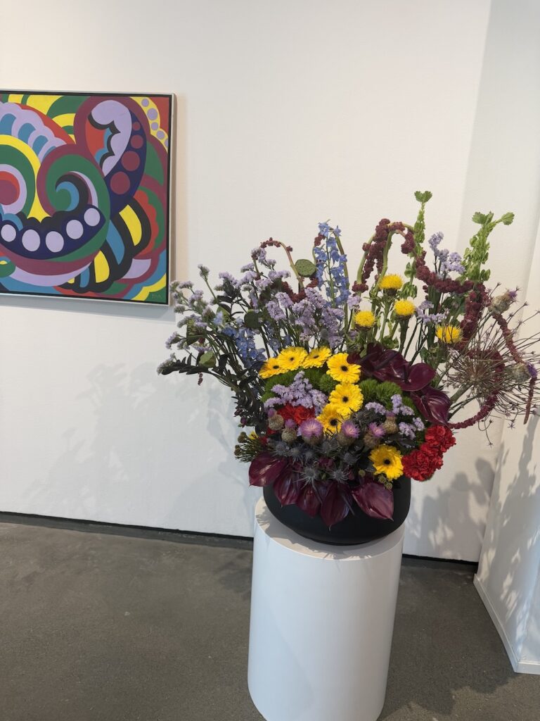 Vibrant abstract painting on the wall beside a tall black vase of mixed flowers on a white pedestal in an art gallery space.