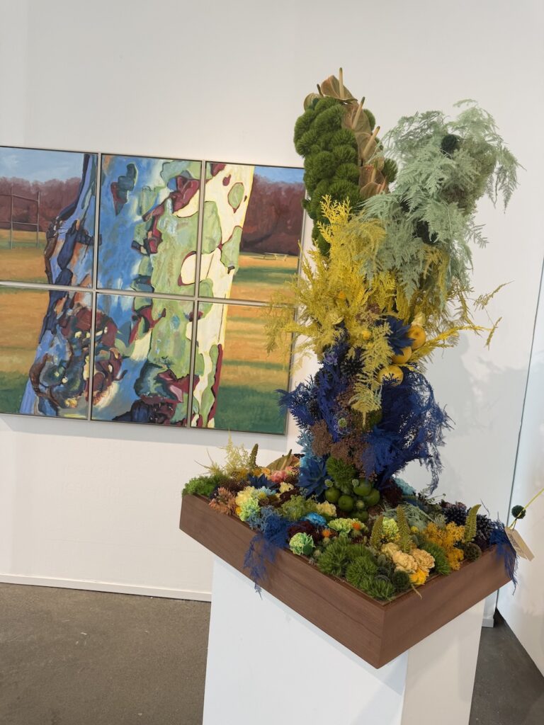 Tall, colorful dried-flower sculpture on a wooden platform in a gallery, with a multi-panel abstract painting in the background.