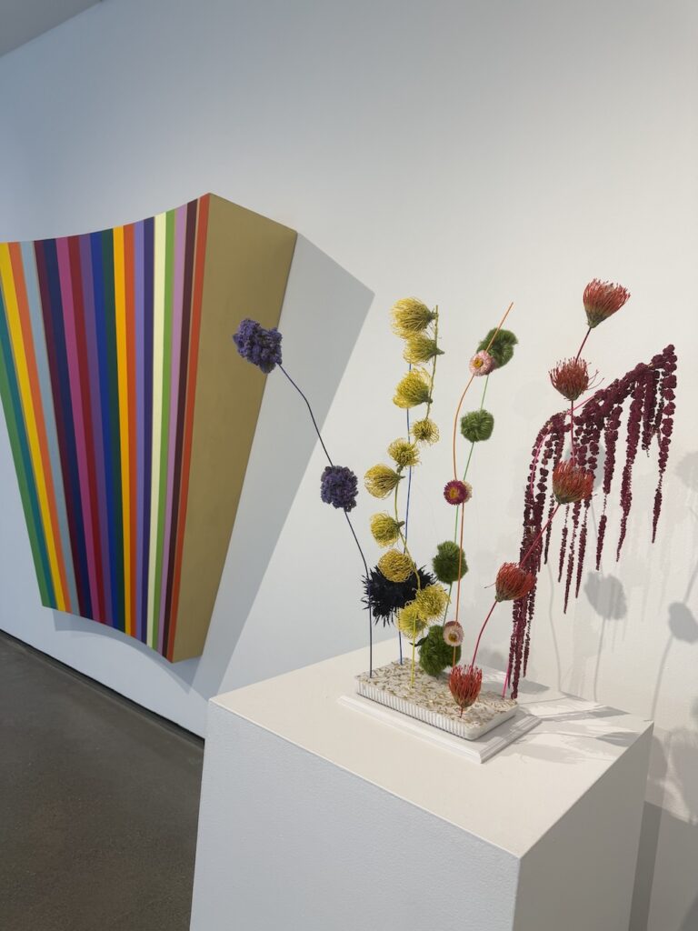 Geometric wall panel with multicolored vertical stripes leaning against a white gallery wall, beside a pedestal with a bright floral sculpture on top.